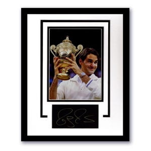 Puede incluir: Fotografía enmarcada de un hombre sosteniendo un trofeo dorado, probablemente un premio deportivo. La foto está sobre un fondo negro e incluye una firma. El marco es negro con un paspartú blanco.