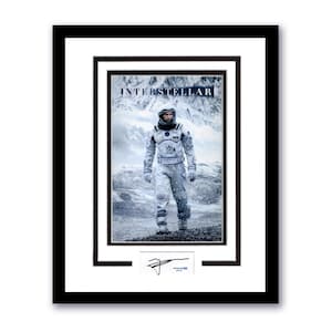 May include: Framed movie poster for the film "Interstellar" featuring a person in a white spacesuit standing in a snowy landscape. The poster has a black frame with a white matting. Includes a signature at the bottom.