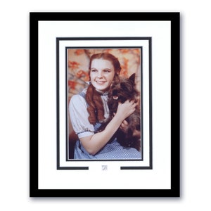 May include: Framed photograph of a woman with red hair, wearing a blue and white checkered dress, holding a small, dark-haired dog. The image is set against a floral backdrop and framed in black with a white mount.