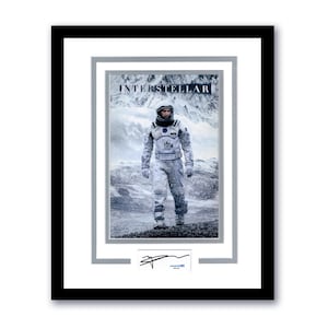 May include: Framed print of the movie "Interstellar" featuring a person in a spacesuit standing in a snowy landscape. The print has a black frame, a white mat, and a signature at the bottom.