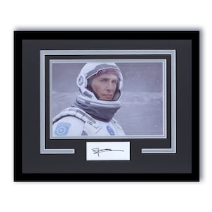 May include: Framed print featuring a man in a white astronaut suit and helmet, set against a snowy backdrop. The print is double-matted with a silver inner mat and a black frame. A white rectangle with a signature is below the image.