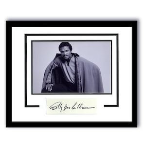 May include: A black and white photograph of a man wearing a cape and a white shirt. The photo is framed in a black frame with a white mount. The man's signature, "Billy Dee Williams", is written in black ink on a white card below the photo.
