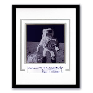 May include: Framed black and white photograph of an astronaut on the moon. The astronaut is wearing a spacesuit and standing on a gray surface. The photograph is signed and includes the text "Hold on to your dreams" and the name "Alan Bean!"