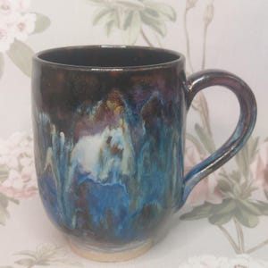 Puede incluir: Una taza de cerámica hecha a mano con un esmalte único. La taza presenta una parte superior marrón oscuro que se transforma en una mezcla de azul, morado y blanco. El asa y la base son de un tono más claro, creando un contraste llamativo. Ideal para café o té.