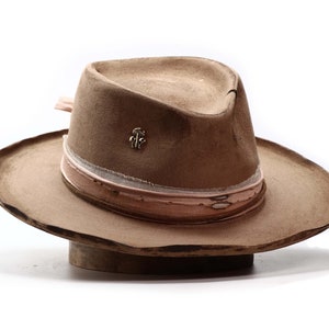 May include: A tan fedora hat with a wide brim. The hat features a light pink and white band around the crown and a small silver-colored pin. The hat is displayed on a wooden hat block against a white background.