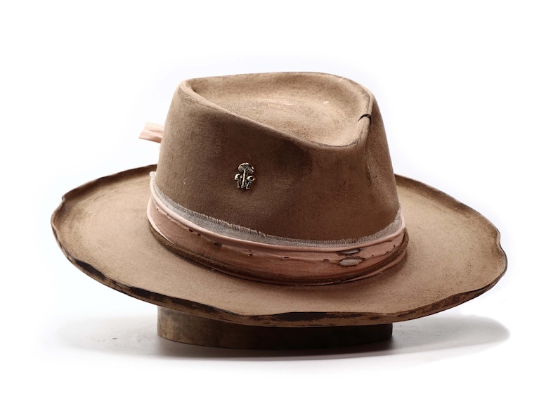 May include: A tan fedora hat with a wide brim. The hat features a light pink and white band around the crown and a small silver-colored pin. The hat is displayed on a wooden hat block against a white background.