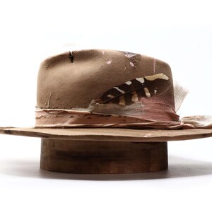 May include: A distressed brown fedora hat with a wide brim. The hat features a pink and cream-colored band, a decorative feather, and a weathered appearance. The hat is displayed on a wooden hat stand.