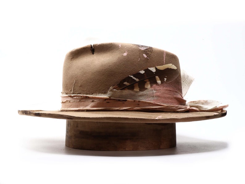 May include: A distressed brown fedora hat with a wide brim. The hat features a pink and cream-colored band, a decorative feather, and a weathered appearance. The hat is displayed on a wooden hat stand.