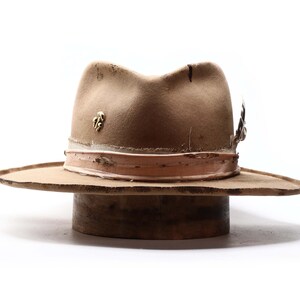 May include: A tan fedora hat with a distressed appearance. The hat features a light brown band, a gold-colored pin, and a feather accent. The hat is displayed on a wooden hat block against a white background.