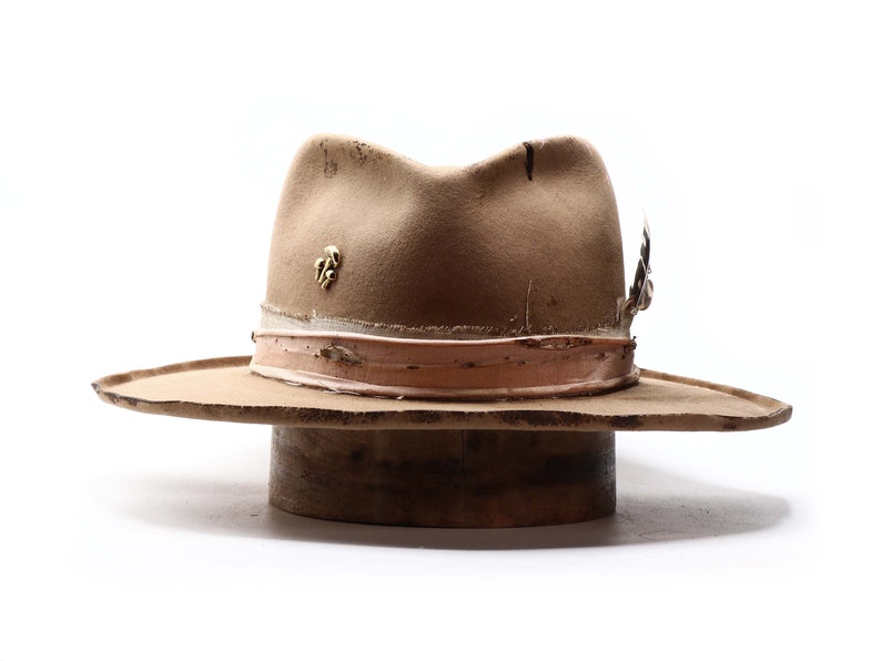 May include: A tan fedora hat with a distressed appearance. The hat features a light brown band, a gold-colored pin, and a feather accent. The hat is displayed on a wooden hat block against a white background.