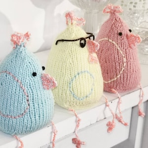 Könnte beinhalten: Drei gestrickte Vogel-Ornamente in Blau, Gelb und Rosa. Jeder Vogel hat ein aufgesticktes Auge und einen kleinen Schnabel. Der blaue Vogel hat einen rosa Schnabel, der gelbe Vogel hat einen rosa Schnabel und eine Brille, und der rosa Vogel hat einen rosa Schnabel.