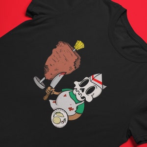 Puede incluir: Una camiseta negra con una ilustraci&oacute;n de dibujos animados de un esqueleto con un gorro de cocinero y un delantal, sosteniendo un cuchillo y un gran trozo de carne. El esqueleto est&aacute; cubierto de salpicaduras de sangre y hay tacos en un plato en la esquina inferior izquierda.