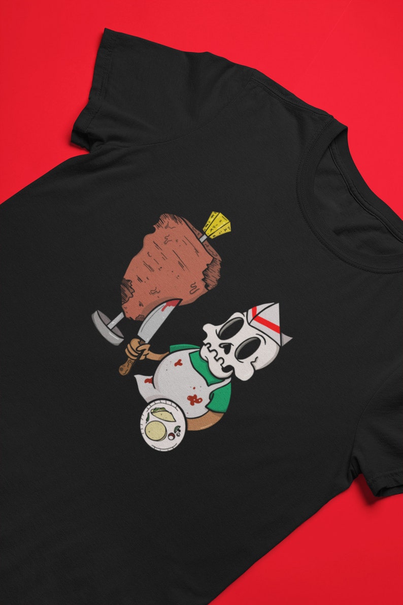 Puede incluir: Una camiseta negra con una ilustraci&oacute;n de dibujos animados de un esqueleto con un gorro de cocinero y un delantal, sosteniendo un cuchillo y un gran trozo de carne. El esqueleto est&aacute; cubierto de salpicaduras de sangre y hay tacos en un plato en la esquina inferior izquierda.