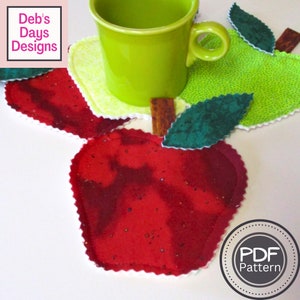 May include: Three red apple-shaped fabric coasters with white scalloped edges. A green apple-shaped coaster with white scalloped edges is on top of the red coasters. A green mug is sitting on top of the green coaster. The coasters have a "PDF Pattern" label.