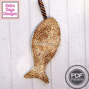 K&ouml;nnte beinhalten: Ein brauner und beiger Stoffbeutel in Fischform mit Leopardenmuster. Der Beutel hat einen braunen und wei&szlig;en gestreiften Stoffgriff. Der Beutel h&auml;ngt an einer wei&szlig;en Holzoberfl&auml;che. Der Text "PDF Pattern" befindet sich auf einem wei&szlig;en Kreis mit grauer Umrandung.