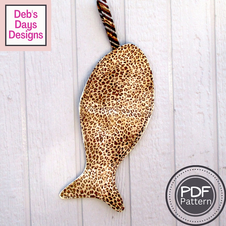 K&ouml;nnte beinhalten: Ein brauner und beiger Stoffbeutel in Fischform mit Leopardenmuster. Der Beutel hat einen braunen und wei&szlig;en gestreiften Stoffgriff. Der Beutel h&auml;ngt an einer wei&szlig;en Holzoberfl&auml;che. Der Text "PDF Pattern" befindet sich auf einem wei&szlig;en Kreis mit grauer Umrandung.