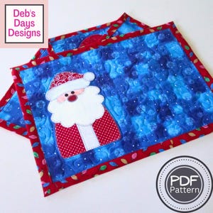 May include: Christmas placemat with a Santa Claus design. The placemat has a blue background with white snowflake accents and a red border. The text "Deb's Days Designs" is in the upper left corner. The PDF pattern is available.