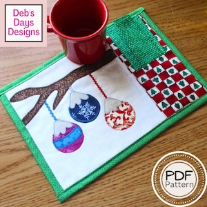 Può includere: Un sottobicchiere in tessuto bianco con bordo verde, che presenta un ramo di albero marrone con tre ornamenti natalizi appesi. Gli ornamenti sono decorati con motivi blu, rossi e bianchi. Il sottobicchiere è su una superficie di legno con una tazza rossa e un quadrato di tessuto verde e rosso.