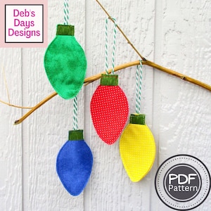 May include: Four colorful felt Christmas light ornaments hanging from a branch. The ornaments are green, red, blue, and yellow, with green tops and white and green string. The red light has white polka dots. The image includes the text "Deb's Days Designs" and a "PDF Pattern" logo.