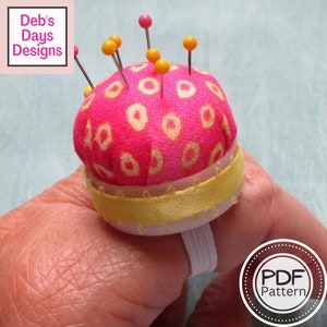 May include: A pink ring pin cushion with yellow and white circular patterns, topped with pins with yellow and pink heads. The cushion has a yellow ribbon trim and a white elastic ring. The text "Deb's Days Designs" and "PDF Pattern" are visible.