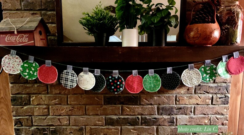 Puede incluir: Una guirnalda navide&ntilde;a hecha de c&iacute;rculos de tela con varios patrones navide&ntilde;os, incluyendo copos de nieve, renos y cuadros. La guirnalda est&aacute; enhebrada con una cinta gris y colgada sobre una chimenea de ladrillo.