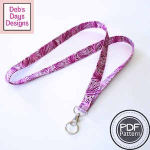 May include: A fabric lanyard featuring a repeating purple and white fan-like leaf pattern. The lanyard includes a silver-toned metal swivel lobster clasp and a circular split ring. The image includes text that reads Deb's Days Designs and PDF Pattern.