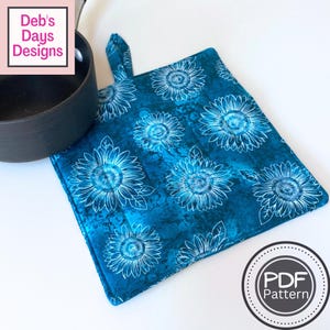 May include: A square fabric potholder featuring a teal and blue marbled background with a white line-art sunflower pattern. The potholder includes a small fabric hanging loop at the top corner and is positioned next to a black metal pot.