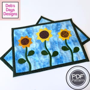 Pode incluir: Duas toalhas de mesa retangulares com fundo azul e três girassóis amarelos. Os girassóis têm centros castanhos e caules e folhas verdes. As toalhas de mesa têm uma borda verde escura. O texto "Deb's Days Designs" está no canto superior esquerdo.
