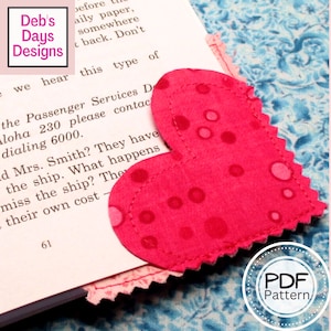 Könnte beinhalten: Ein rosafarbenes herzförmiges Stoff-Lesezeichen mit weißen Punkten. Das Lesezeichen liegt auf einem Buch mit dem Text "Deb's Days Designs" und "PDF Pattern".