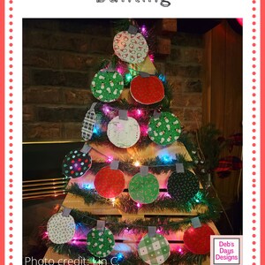 Puede incluir: Una guirnalda de adornos navide&ntilde;os hecha de c&iacute;rculos de tela con varios patrones festivos, incluyendo mu&ntilde;ecos de nieve, bastones de caramelo y cuadros. La guirnalda est&aacute; enhebrada en un marco de madera y decorada con una cadena de luces. La imagen incluye el texto "Deb's Days Designs" y "www.debsdays.com".