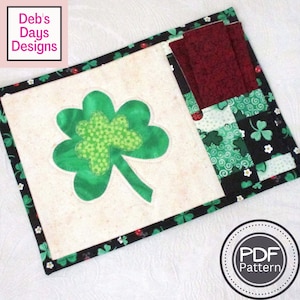 May include: A green shamrock-shaped fabric applique on a white background with a black and green fabric border. The fabric border has a pattern of shamrocks, swirls, and ladybugs. A red fabric pocket is on the right side of the mat. The text "PDF Pattern" is on a white circle with a grey border.