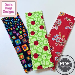May include: Three rectangular fabric bookmarks with pinked edges. One features a black background with colorful squares, one has a light green background with red ladybugs and leaves, and one has a red background with floral patterns. Text reads Deb's Days Designs.