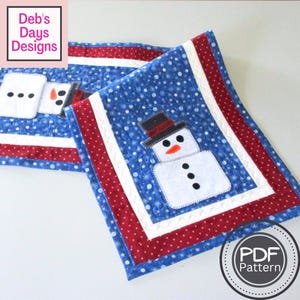 May include: Two blue quilted placemats with a snowman design. The placemats have a white border, red polka dot trim, and a snowman applique with a black top hat, orange nose, and black button eyes and mouth. The text "Deb's Days Designs" is in the upper left corner.