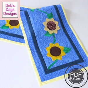 May include: A quilted table runner featuring a blue background with a sunflower design. The sunflowers have yellow petals and brown centers. The runner has a yellow border and the text "Deb's Days Designs". A PDF pattern is available.