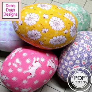 May include: Five fabric Easter eggs in various colours and patterns. The eggs are decorated with floral prints, polka dots, and bunny silhouettes. The eggs are arranged in a cluster on a checkered surface. The text "PDF Pattern" is visible in the lower right corner of the image.