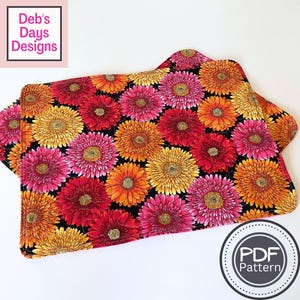 May include: A rectangular fabric placemat featuring a vibrant floral pattern of pink, orange, and yellow gerbera daisies on a black background. The edges are rounded and stitched. The image includes text reading Deb's Days Designs and PDF Pattern.