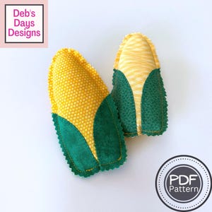 May include: Two plush corn cobs. The corn kernels are yellow with a grid pattern, and the husks are green. The image includes the text "Deb's Days Designs" and a "PDF Pattern" logo.