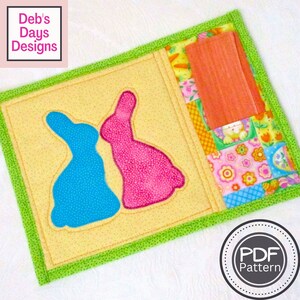 May include: A PDF pattern for a quilted Easter bunny coaster with a pocket. The coaster is yellow with green trim and features two bunnies, one blue and one pink. The pocket is made of colorful patchwork fabric.