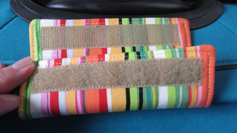 May include: Two colorful striped fabric luggage straps with velcro closures. The straps are attached to a blue suitcase.