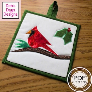 May include: A red cardinal bird perched on a branch with green holly leaves. The bird is made of fabric and is part of a quilted pot holder. The pot holder has a green border and a loop for hanging. The text "PDF Pattern" is visible in the lower right corner of the image.