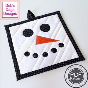 Puede incluir: Un posavasos acolchado blanco con un borde negro de lunares. El posavasos tiene la forma de la cara de un muñeco de nieve con ojos negros, una nariz de zanahoria y una fila de botones negros.