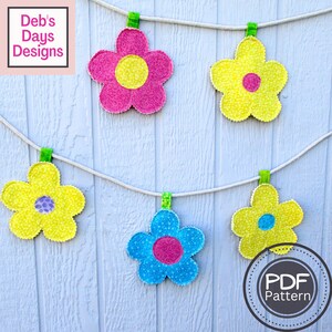 May include: A string of five fabric flower decorations hanging from a white rope with clothespins. The flowers are yellow, pink, blue, and have a smaller circle in the center. The flowers are hanging on a white wooden background. The text "PDF Pattern" is in a circle at the bottom right of the image.