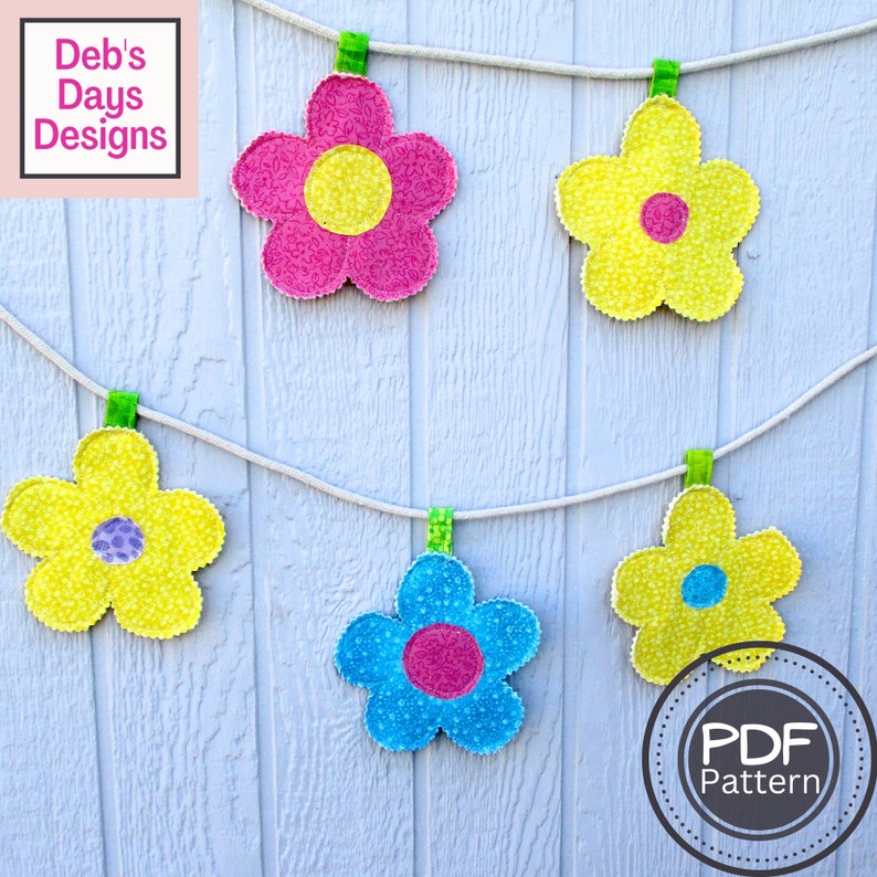 May include: A string of five fabric flower decorations hanging from a white rope with clothespins. The flowers are yellow, pink, blue, and have a smaller circle in the center. The flowers are hanging on a white wooden background. The text "PDF Pattern" is in a circle at the bottom right of the image.