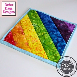 May include: Rainbow-coloured fabric pot holder with a light blue border. The fabric has a batik pattern with floral designs. The pot holder is made of quilted fabric. The text "PDF Pattern" is in a circle on the bottom right corner of the image.