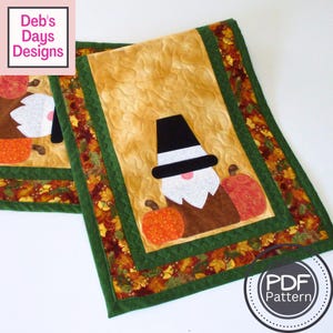 以下が含まれることがあります： 秋をテーマにしたキルトのテーブルランナー2枚。各ランナーは、黒い帽子、白いひげ、オレンジ色のカボチャを持つノームが特徴です。ボーダーは緑色のトリムと秋の葉のパターンです。「Deb's Days Designs」の文字が左上にあります。