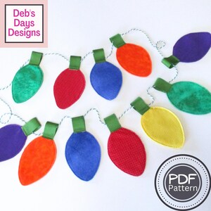 May include: A string of colorful, felt Christmas light ornaments in various colors, including red, blue, orange, green, yellow, and purple. Each light has a green ribbon and is strung on a white and green cord. The image includes the text "Deb's Days Designs" and "PDF Pattern".