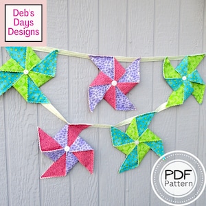May include: A garland of colorful fabric pinwheels in shades of pink, purple, green, and turquoise. Each pinwheel is attached to a light green ribbon. The image includes the text "Deb's Days Designs" and "PDF Pattern".