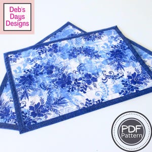 May include: Two rectangular placemats with a blue floral pattern on a white background. The placemats have a dark blue border and are quilted. The text "Deb's Days Designs" is in the upper left corner. A "PDF Pattern" logo is in the lower right corner.