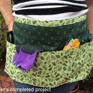 May include: A green gardening apron with multiple pockets, featuring a leafy print. The apron has a green pocket with a seed packet that says "Poppy" and a purple and black pair of gardening gloves.