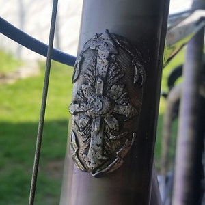 May include: Close-up of a silver bicycle frame with a decorative metal emblem featuring a stylized floral design.